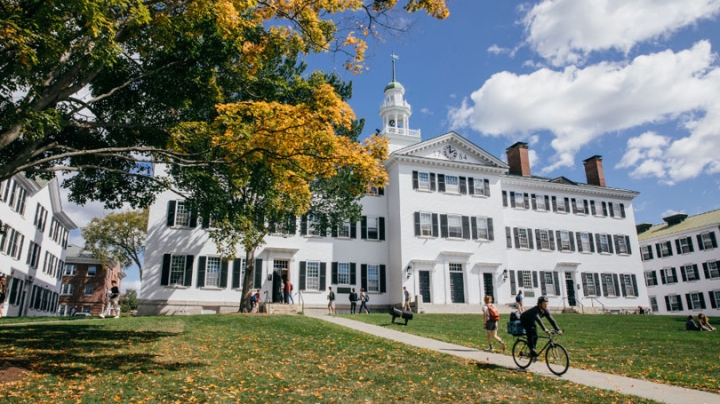 Dartmouth Hall during the fall. Students are walking to and from the hall and there is one riding his bike away from the hall. There is a tree with bright golden leaves. 